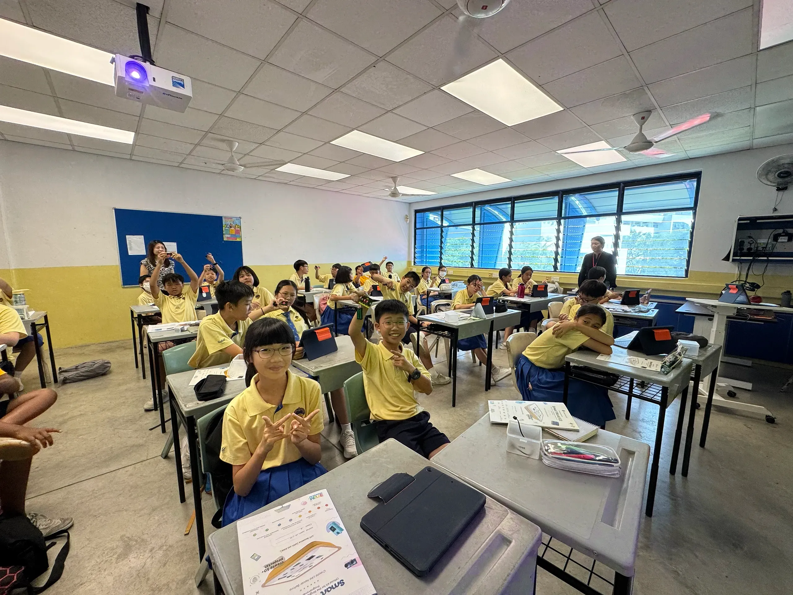 A class of students in school uniforms is actively engaged in a technology-enhanced lesson. Seated at individual desks equipped with tablets and learning materials, many students raise their hands to answer questions or share ideas. The bright classroom features a projector, large windows, and a teacher facilitating the session, creating a collaborative and interactive learning environment.